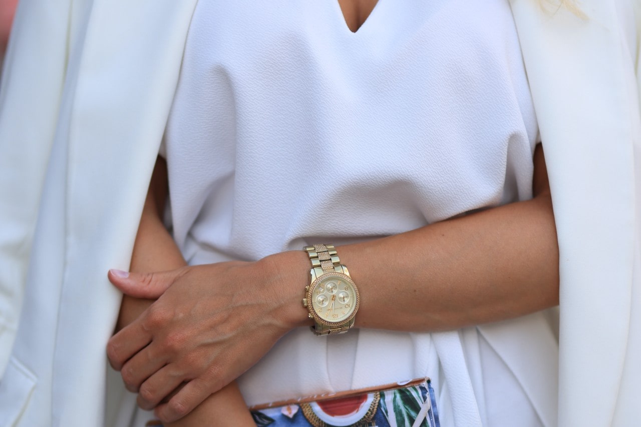 Close up of a woman wearing all white with a gold watch on her wrist and holding a colorful clutch in the other hand
