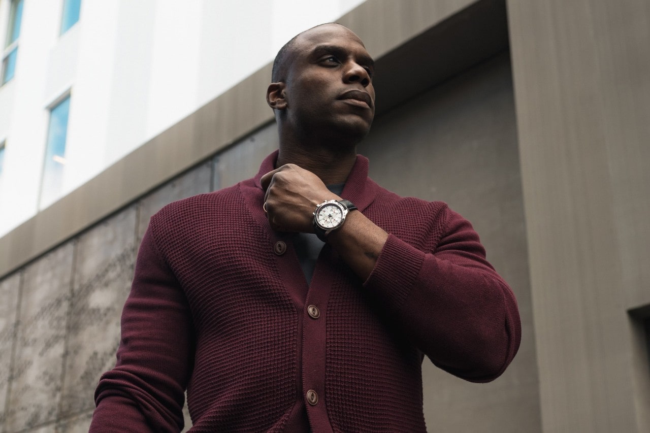 A man in a maroon sweater lifting his hand to his chin, exposing a silver watch with white dial and black leather strap
