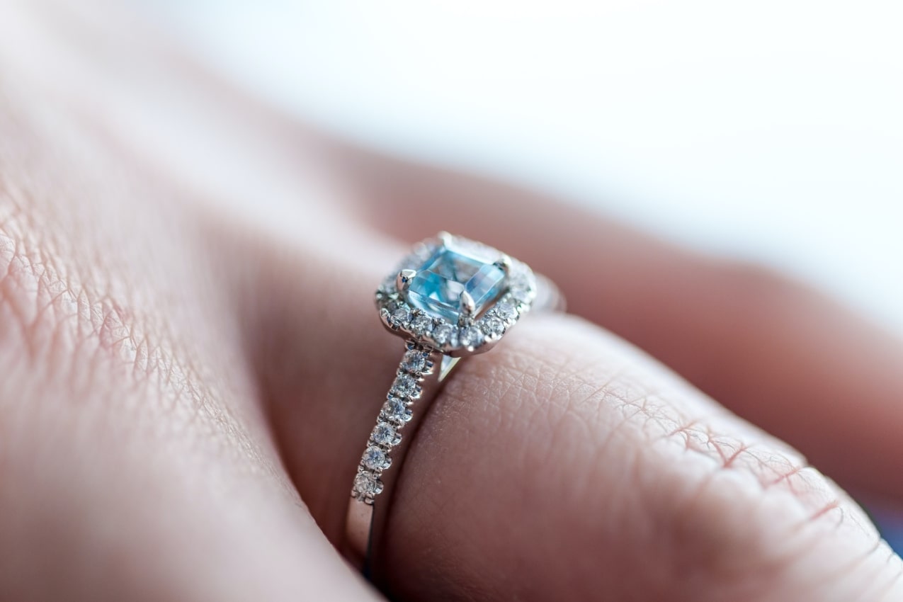 Close up image of a person’s ring finger adorned with an engagement ring featuring a light blue stone