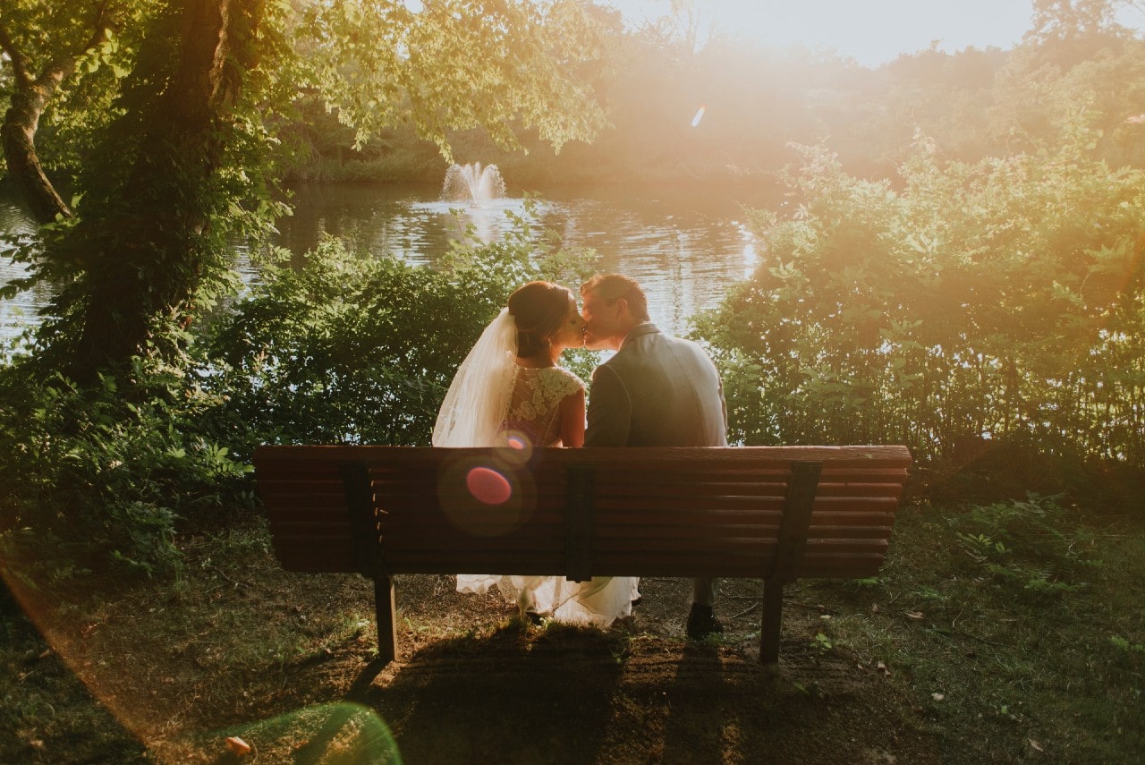 Bride and groom kiss on a park bench beside a lake in July