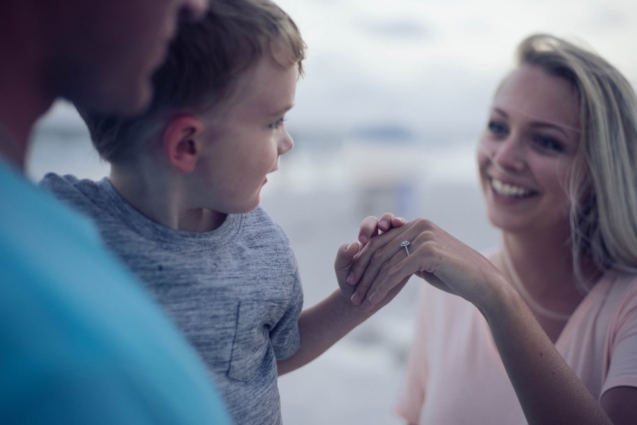 A blonde woman shows her engagement ring to her son while walking on a beach