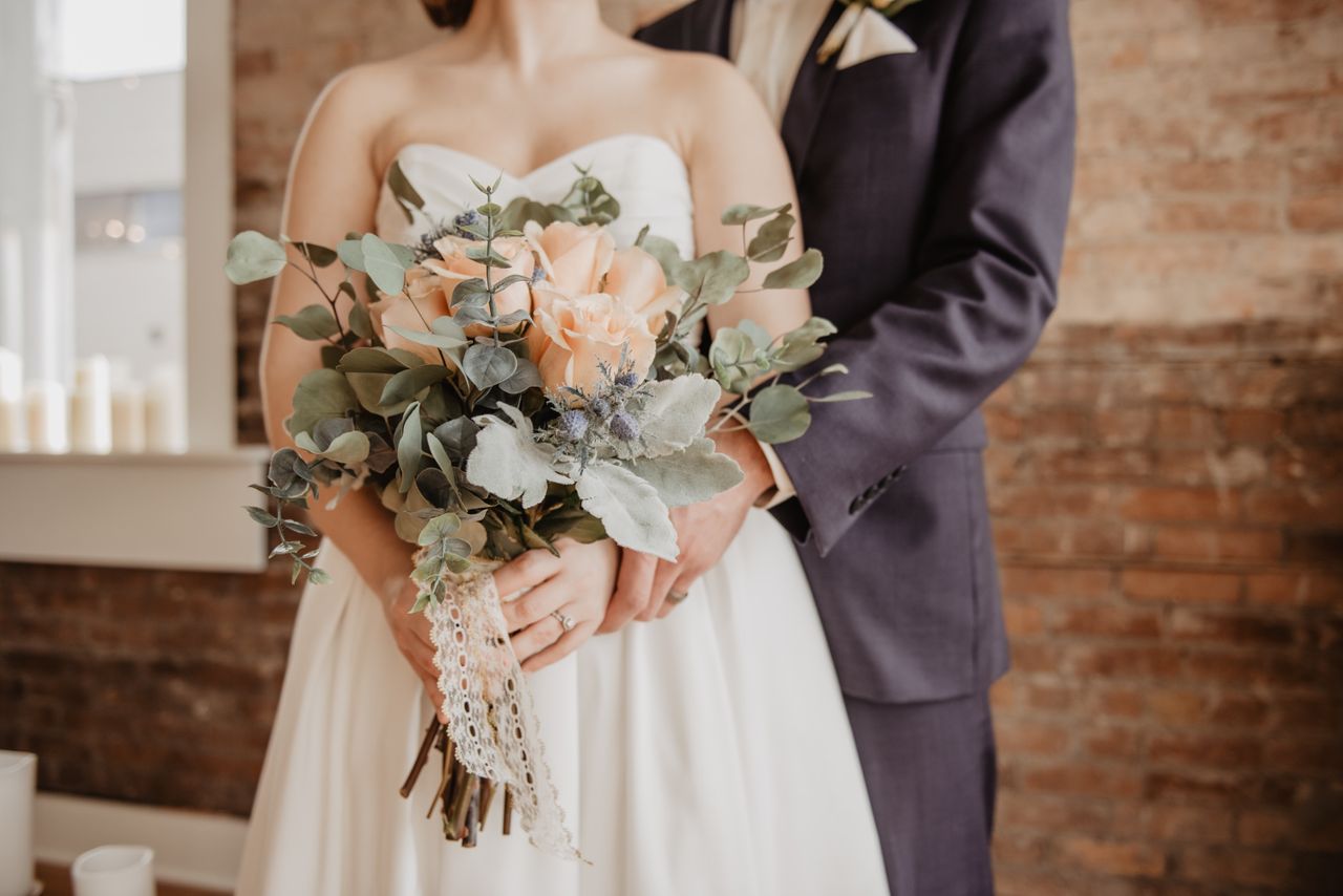A faceless groom and bride stand beside each other while the bride holds her rose bouquet