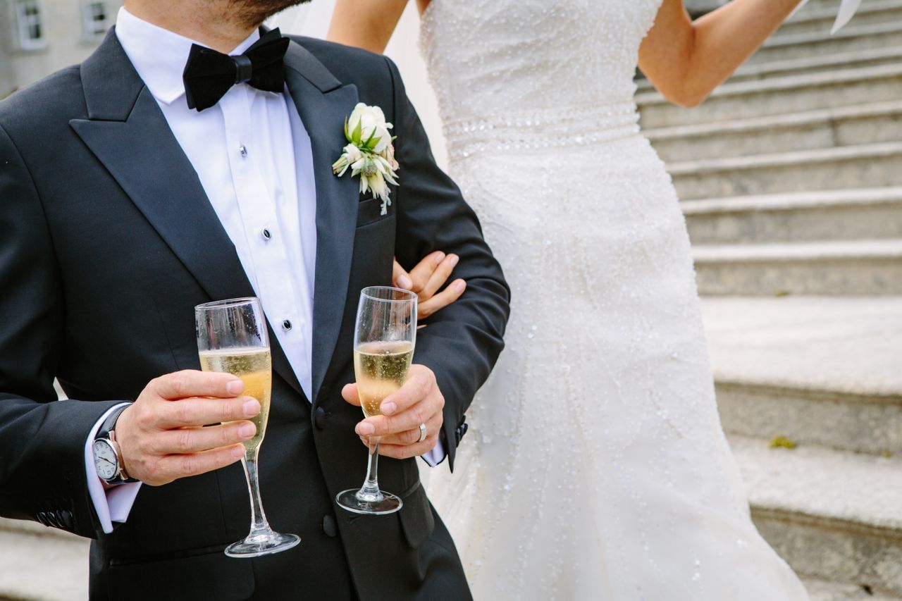 A bride and groom share drinks during their reception. The groom holds two flutes of champagne for both of them.