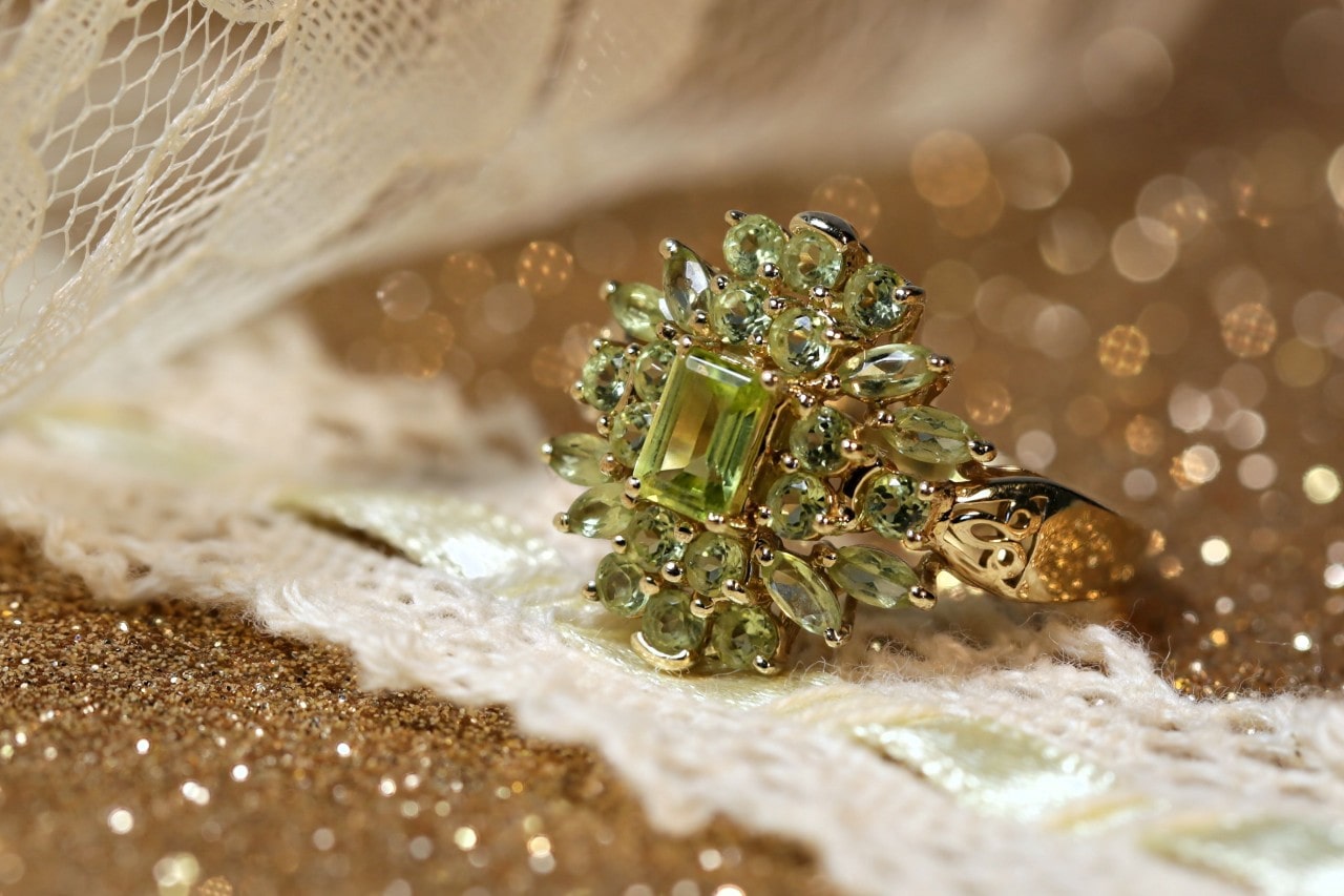 A gold halo peridot ring sits on a gold glittery surface and lace