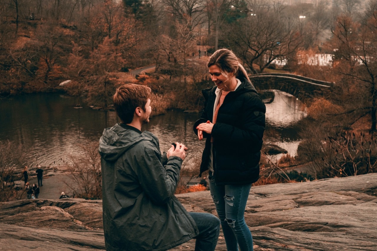 A man down on one knee, presenting a ring to a woman in front of a river in the fall