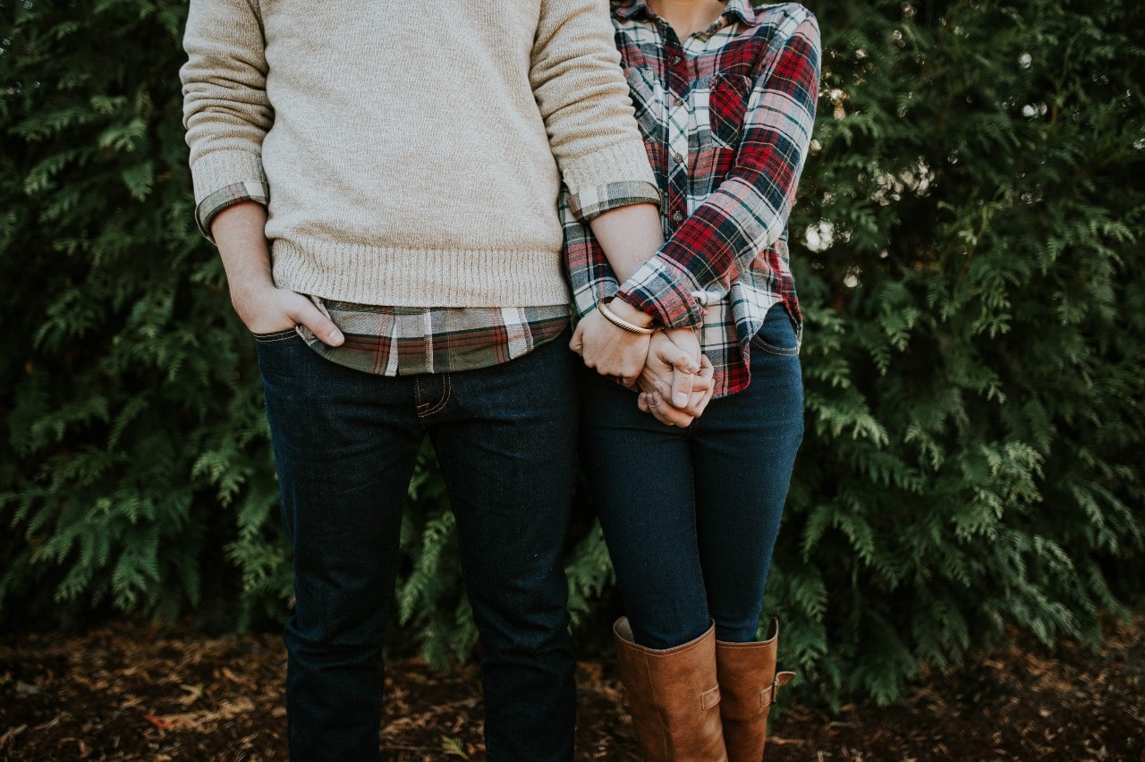 A couple holding hands and wearing plaid in front of pine trees in the fall